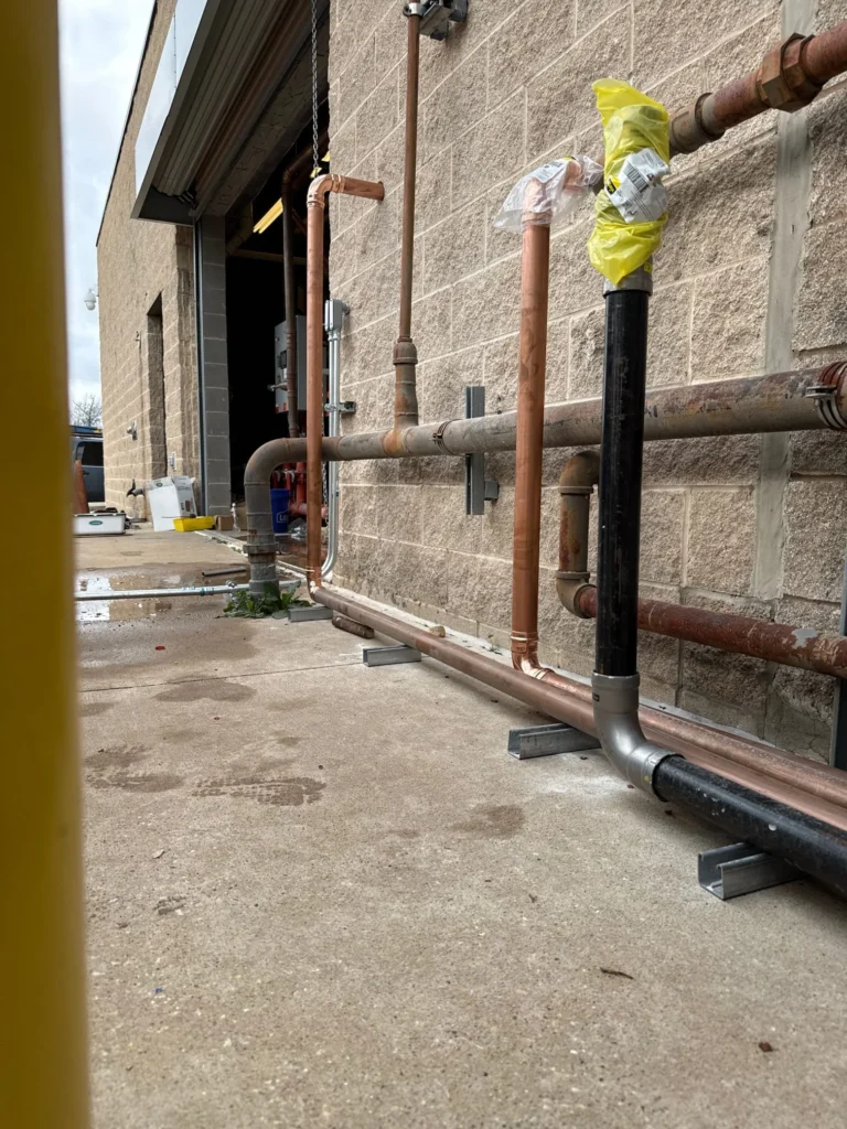 Outdoor plumbing installation with copper and black pipes secured along a brick wall, some wrapped in protective plastic.
