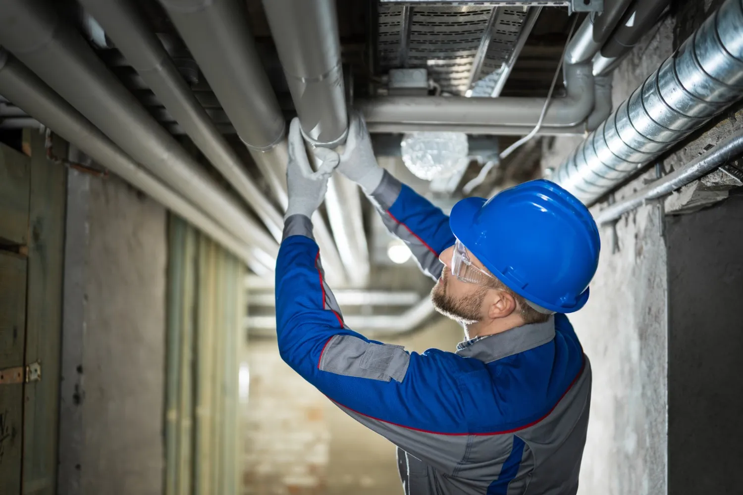 A plumber wearing a blue hard hat safety glasses and blue and grey workwear is looking up and adjusting grey pipes on a ceiling in what appears to be a commercial building