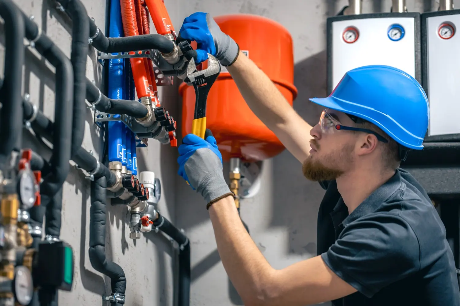 A plumber wearing a blue hard hat safety glasses and blue gloves is using a wrench to work on a system of red and blue pipes and valves in a commercial setting