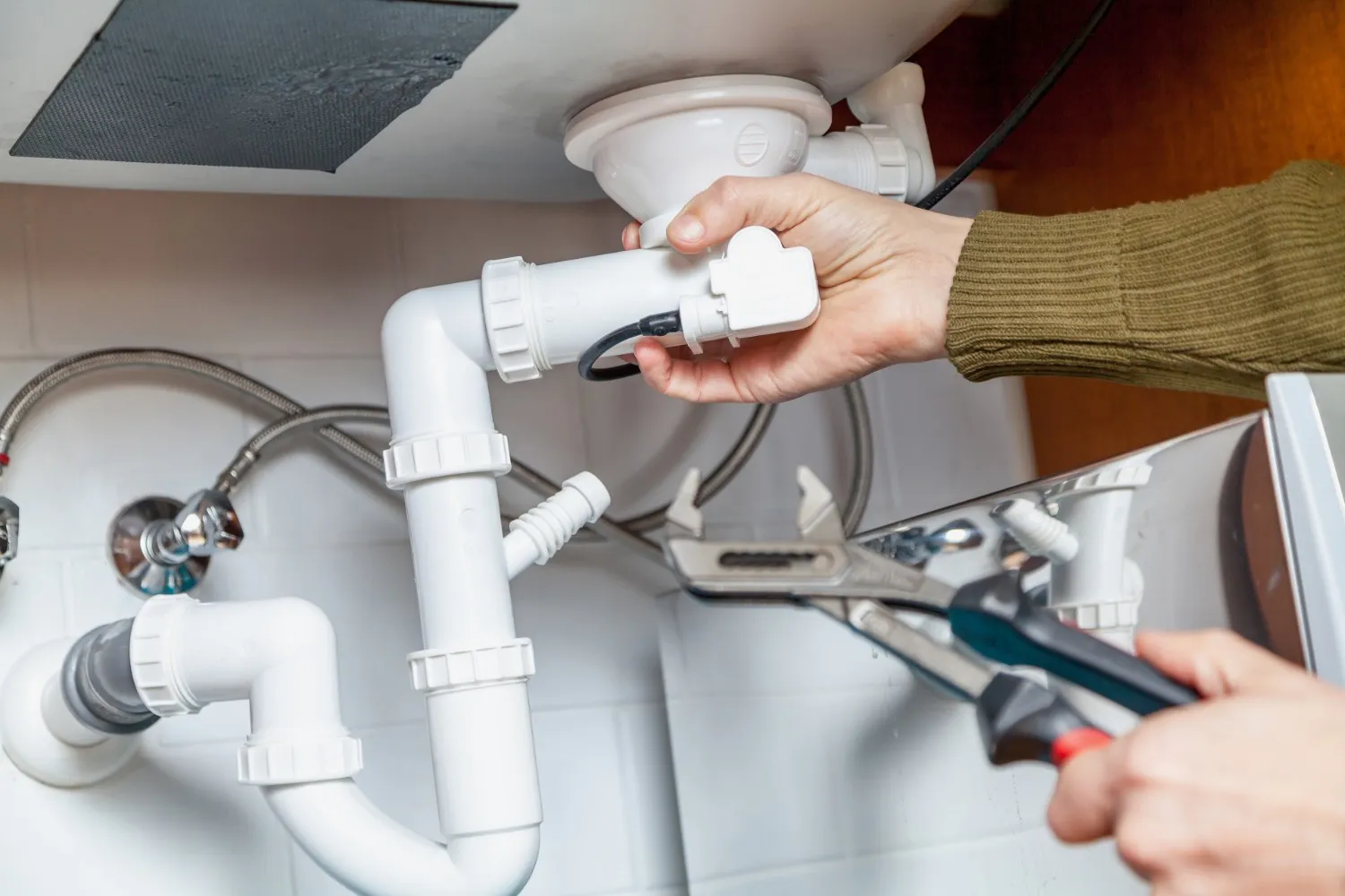 A close-up of a person's hands using a wrench to tighten or loosen a pipe fitting, possibly under a sink, with tools and plumbing components visible.