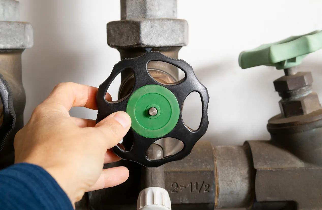 Backflow Prevention in Livingston, TX | McWilliams Heating, Cooling and Plumbing Hand turning black valve wheel with green center on industrial pipe system labeled '4-1/2', featuring green-handled valve and metal fittings