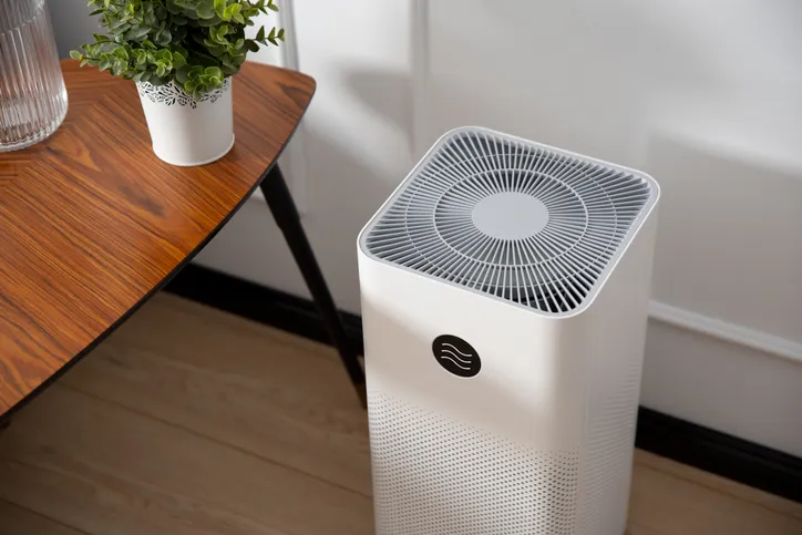 White cylindrical air purifier with top vent on wooden floor beside table with potted plant
