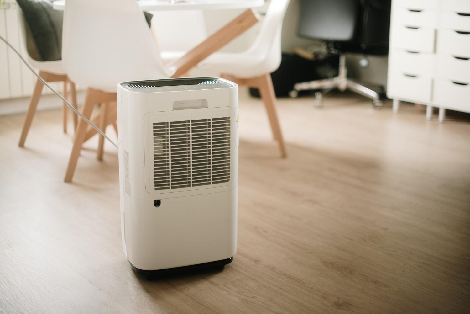 A white rectangular dehumidifier with a large front grille sits on a light wooden floor In the blurred background a dining area with a white table and modern white chairs is visible along with a white storage cabinet