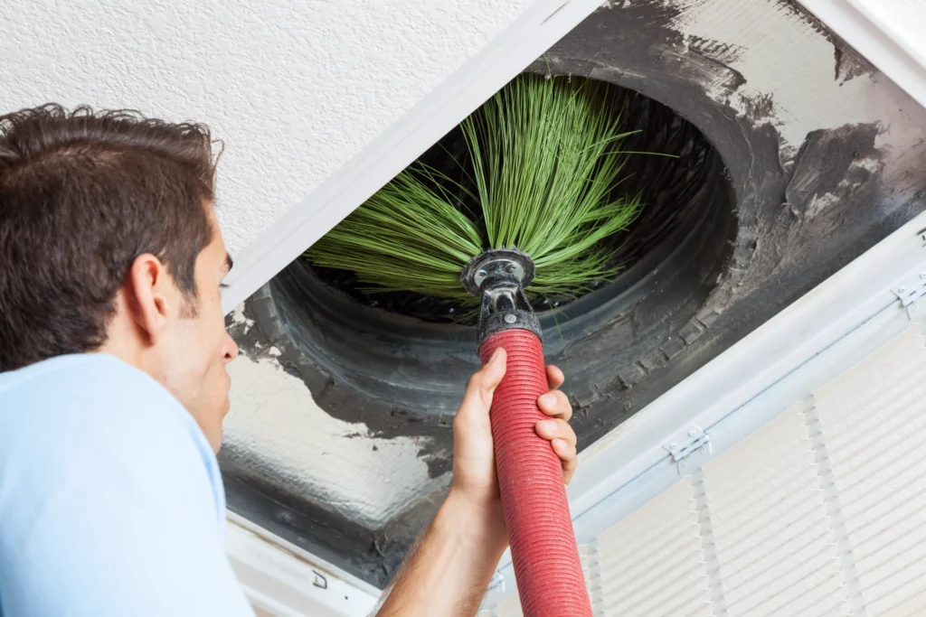A male technician in a light blue polo shirt is performing air duct cleaning inserting a large corrugated red hose into a rectangular vent opening in a white ceiling A ceiling fan and windows with curtains are visible in the background