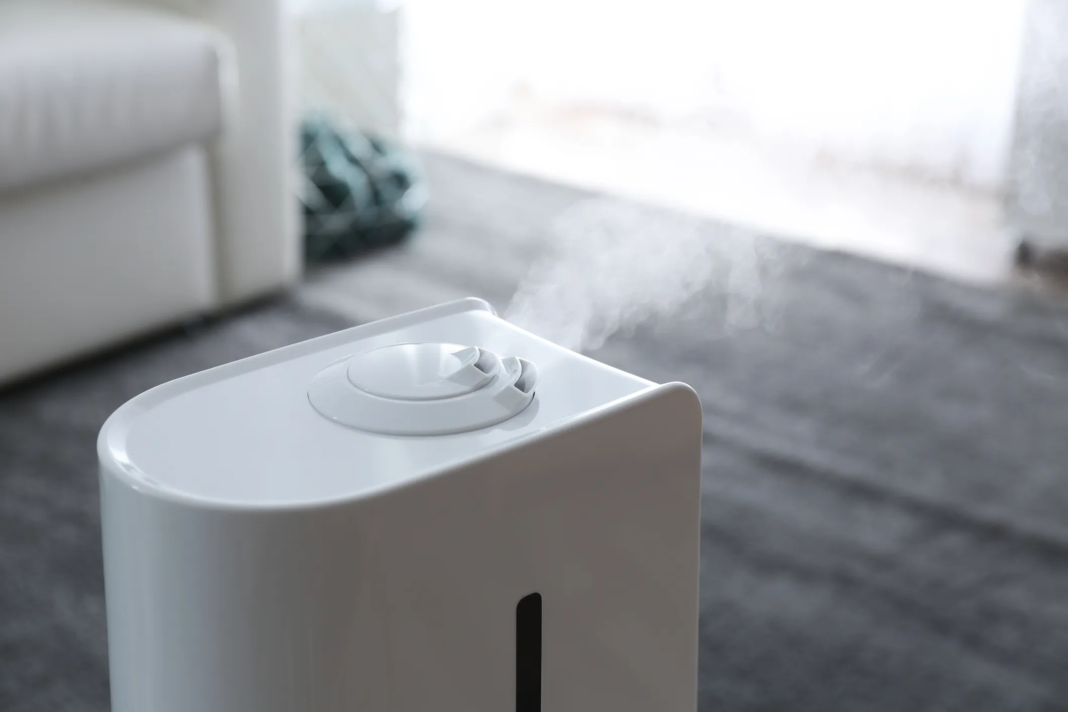 White humidifier emitting mist on dark carpeted floor near white sofa and green fabric