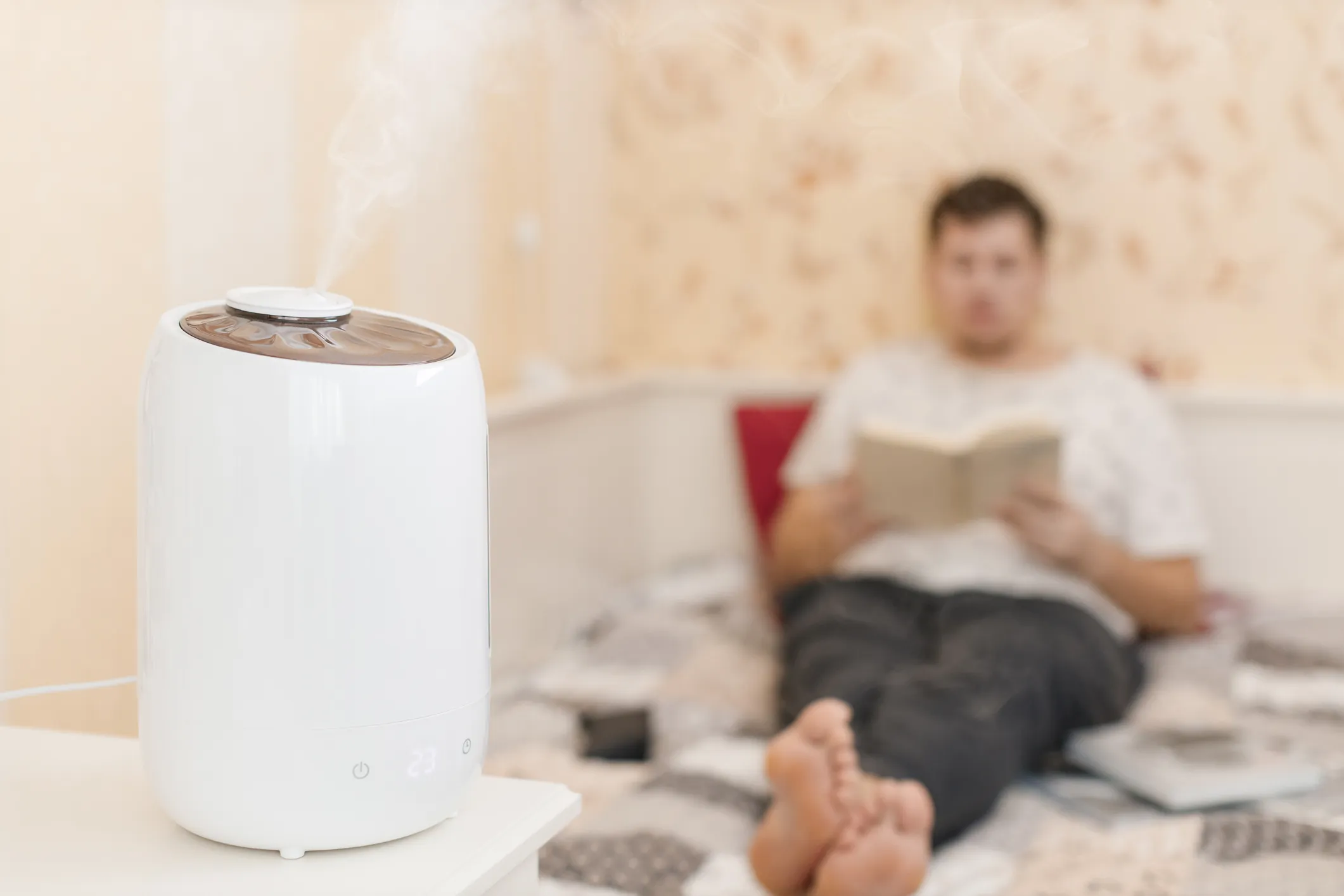 White humidifier emitting mist on bedside table with person reading on bed in cozy room